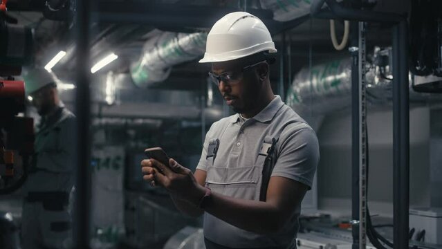 African American engineer uses mobile phone to inspect pipeline system on plant. Professional male worker in safety uniform and protective hard hat works on modern factory or industrial facility.