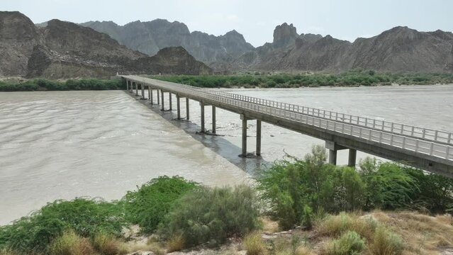 Aerial drone shot over a road bridge over a river flowing with mountains in the background in Hingol, Balochistan, Pakistan on a cloudy day.
