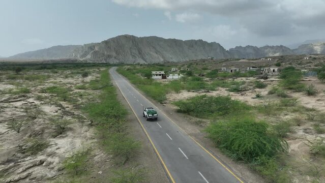 Aerial drone shot over a car driving beside village with mountain range in the background in Hingol, balochistan, Pakistan on a cloudy day,