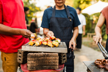 Skewers with colorful vegetables and meat grilling, family and friends gathering in the background, enjoying a sunny outdoor meal. BBQ party, barbeque grill weekend, happy family.