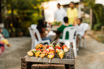 Skewers with colorful vegetables and meat grilling, family and friends gathering in the background, enjoying a sunny outdoor meal. BBQ party, barbeque grill weekend, happy family.