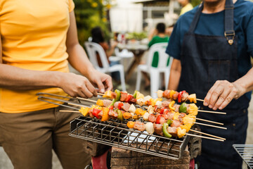 Skewers with colorful vegetables and meat grilling, family and friends gathering in the background, enjoying a sunny outdoor meal. BBQ party, barbeque grill weekend, happy family.