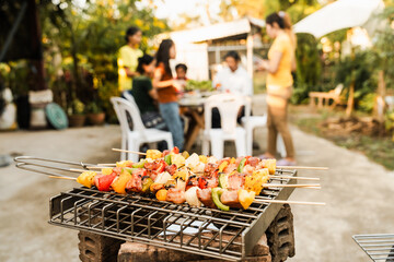 Skewers with colorful vegetables and meat grilling, family and friends gathering in the background, enjoying a sunny outdoor meal. BBQ party, barbeque grill weekend, happy family.