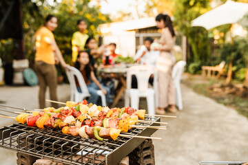 Skewers with colorful vegetables and meat grilling, family and friends gathering in the background, enjoying a sunny outdoor meal. BBQ party, barbeque grill weekend, happy family.