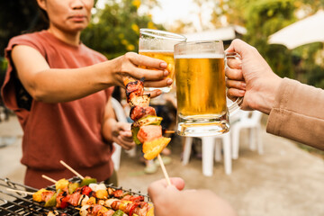 Outdoor barbecue with various meats on grill, two people holding plates and drinks, smoke rising, festive and casual atmosphere. holding glass of beer. family party.