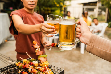 Outdoor barbecue with various meats on grill, two people holding plates and drinks, smoke rising,...