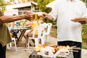 Outdoor barbecue with various meats on grill, two people holding plates and drinks, smoke rising, festive and casual atmosphere. holding glass of beer. family party.