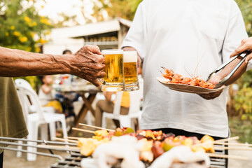 Outdoor barbecue with various meats on grill, two people holding plates and drinks, smoke rising, festive and casual atmosphere. holding glass of beer. family party.