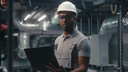 Male African American inspector checks piping system using laptop and looks at camera. Diverse team of professional technician specialists work on modern factory. Heavy industry concept. Portrait.