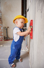 Child construction worker checking wall surface with spirit level tool. Full length of kid in work overalls using level instrument while preparing wall for repair work at home during renovation.