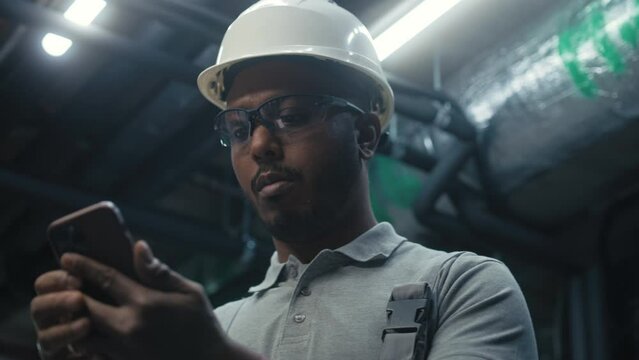 Close up of African American engineer using phone during break, surfing the Internet. Heavy industry worker in safety uniform and hard hat works on modern manufacturing factory or industrial plant.