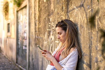 Obraz premium Engrossed in Technology: A Side-View, Horizontal Shot of an Unidentified Individual Leaning Against a Wall, Engaged in Texting on Their Phone, Under the Warm Sunlight of the Day