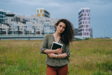 Pensive young woman holds tablet stands at meadow against view on new building on background. Professional architect planning new block.
