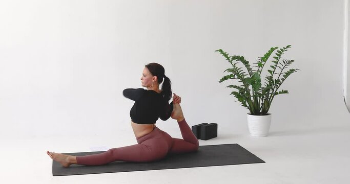 Yoga instructor performing Hanumanasana exercise with ring grip, training in leggings and long sleeve top while sitting on mat in studio
