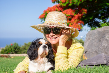 Happy senior smiling woman in hat and sunglasses lying in the meadow with her cavalier king charles...