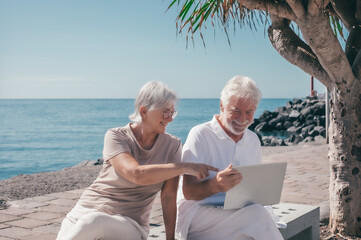 Cheerful modern senior couple sitting on a bench close to the sea using laptop connection together...