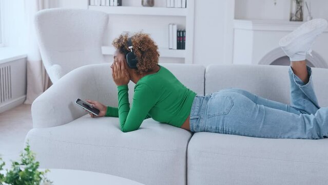 Young beautiful casual African American woman zoomer lying on sofa listening to music on headphones using mobile phone with new audio application for melomans located in bright stylish living room.
