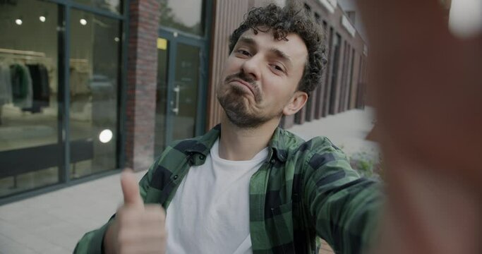 Slow motion portrait of happy Middle Eastern man taking selfie posing looking at camera showing thumbs-up gesture in street. Photo and urban lifestyle concept.