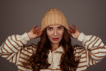 A beautiful girl of European appearance in a beige large-knit hat and sweater on a gray background holding on to the hat