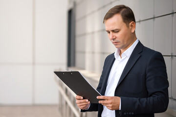Concentrated mature businessman dressed in suit stands near office reading contract. Adult concerned man learns documents with interest