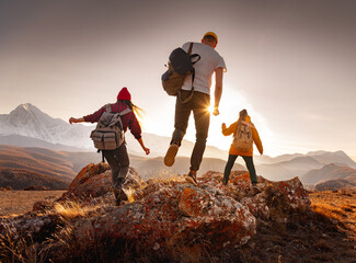 Three young hikers with small backpacks walks at sunset mountains. Weekend activities concept - obrazy, fototapety, plakaty