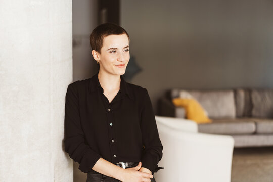 Casual Elegance: Smiling Young Woman with Short Hair Leaning Against Wall in Modern Living Room