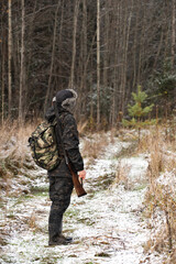 Male hunter in camouflage and with backpack, armed with a rifle, walks through the snowy winter forest