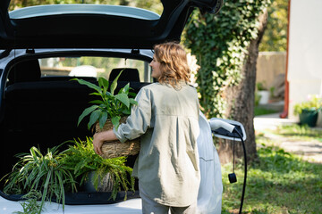 Woman with plant in pot next to a charging electric car in the yard of a country house.
