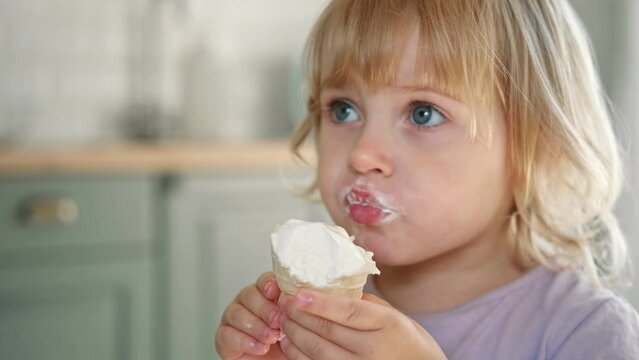 Baby girl enjoying ice cream. Pretty little toddler eating an ice-cream indoors, at home. Dining room background. Small child eats plombir and cream messy on her mouth. Cute kid with tasty sweet food.