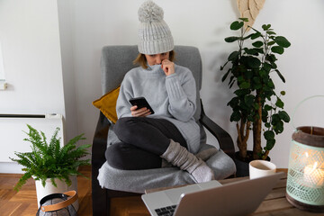 A woman in warm clothes with smartphone sits close to a heating radiator. Low temperature in the...