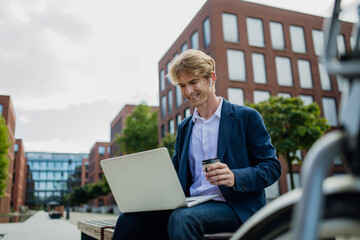 Businessman, freelancer or manager working outdoors in city park. Man with laptop on knees drinking coffee, having video call, listening music. Concept of working remotely.