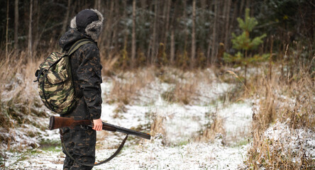 Male hunter in camouflage and with backpack, armed with a rifle, walks through the snowy winter forest