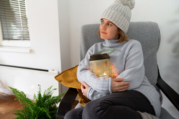 A woman in warm clothes sits in an armchair and hugs a lantern with candle. Low temperature in the...