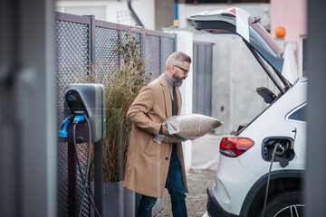 Man putting bag of wood pellets in car trunk, while his electric vehicle is charging.