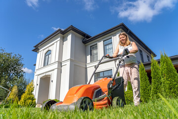 Pregnant woman mows green lawn in the backyard near country house, cleaning territory with  electric lawn mower for cutting grass on back yard on sunny summer day