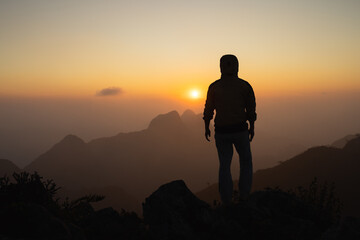 Silhouette of a hiker man on a rock pedestal with hands up. Beautiful orange sunset. Independent...