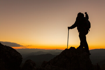 Silhouette of a hiker man on a rock pedestal with hands up. Beautiful orange sunset. Independent hiking travel, success.