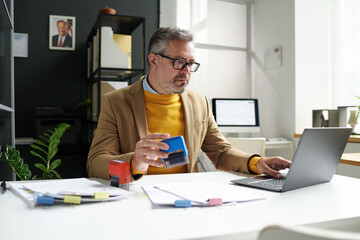 Mature manager sitting at his workplace and putting stamps on documents in office