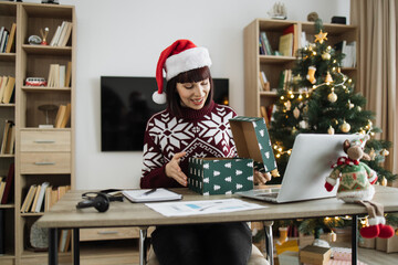Excited Caucasian woman in Santa hat unpacking gift while sitting at desk with laptop. Happy female opening wrapped box and feeling excited during remote work at home decorated with Christmas tree.