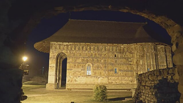 Night view of Humor Monastery with its illuminated painted walls, framed by stone arch, famous UNESCO heritage in Bukovina, Romania