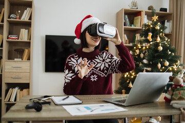 Caucasian young adult businesswoman wearing Santa hat using VR glasses touching air and gesturing, sitting at table with gadgets at home near xmas tree during the holiday season.