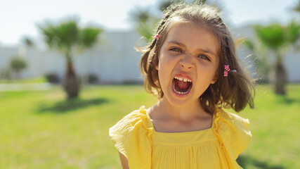 portrait of girl 7years old in yellow dress screams at camera on lawn with palm trees in summer day