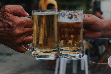 two hands holding frosty mugs of beer, one full and one half-empty, during what appears to be a social event.
