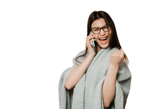 Excited young girl clenches fist like winner  talks by phone received great news, wrapped in knitted blanket against transparent background, looks at camera. Young in glasses celebrating success