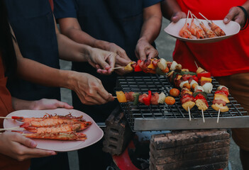 People are cooking seafood and vegetable skewers on a charcoal grill. with a plate of shrimp placed...
