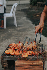People are cooking seafood and vegetable skewers on a charcoal grill. with a plate of shrimp placed...