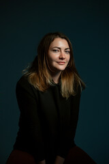 Classic dark studio portrait of a young brown-haired woman in black clothes sitting on a chair.
