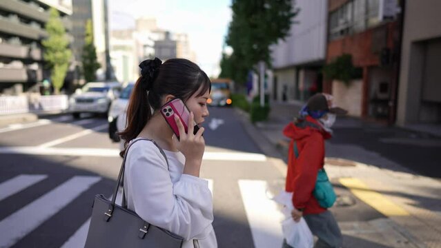 愛知県名古屋市大須商店街周辺の道路沿いでスマートフォンで通話しながら歩く若い日本人女性 Young Japanese woman walking while talking on her smartphone along a road near the Osu shopping district in Nagoya City, Aichi Prefecture, Japan.