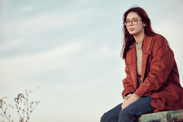 Pensive calm teenage girl in glasses with long red hair in red coat sits on concrete podium and looks away, against the background of sky .