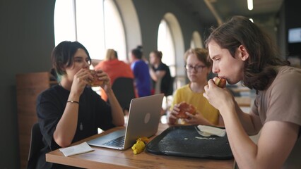 group of students sit in a cafe and do a project on a laptop together. business concept of modern training and lifestyle development. students discuss their homework and eat fast food burgers